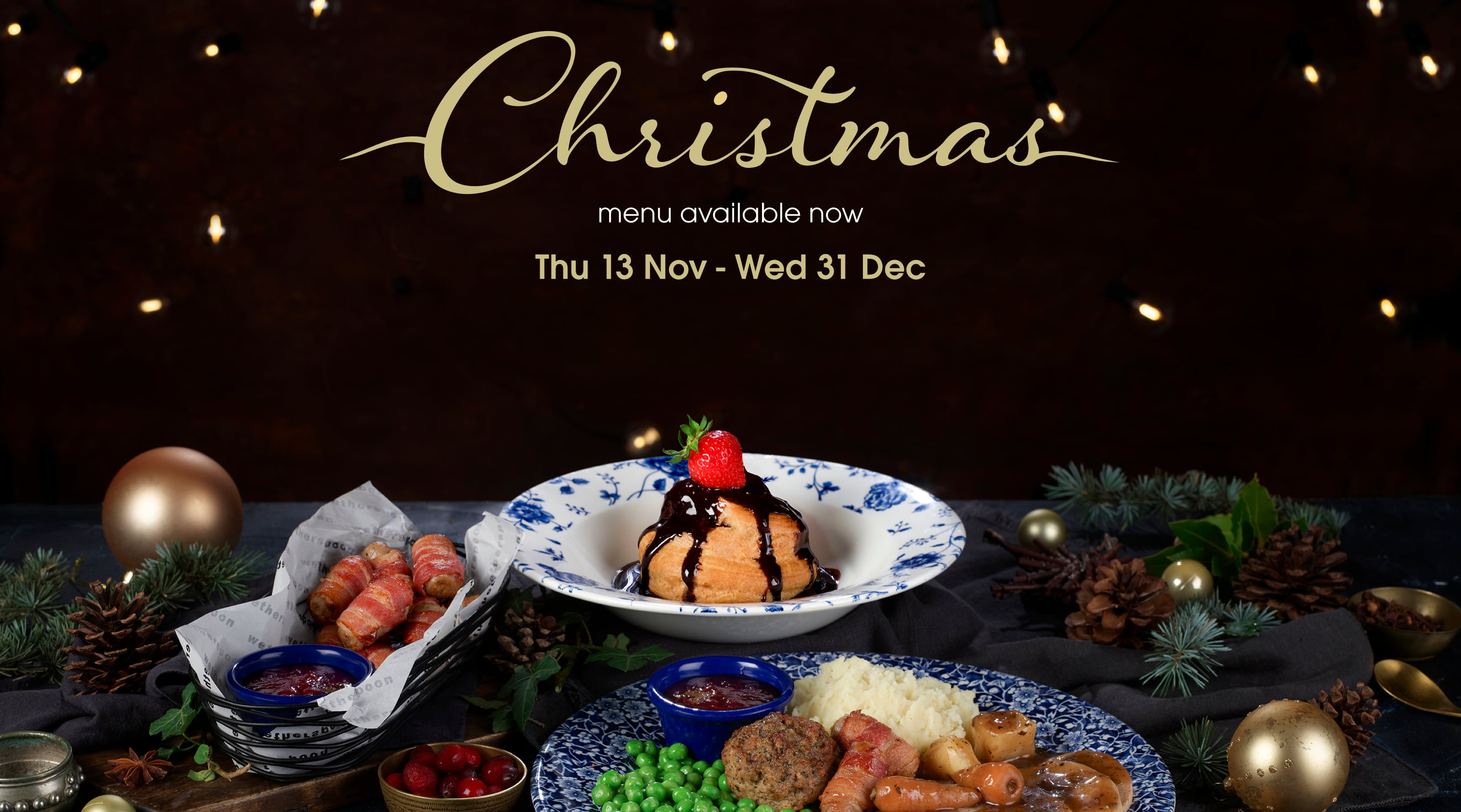 Artistic shot of a chocolate Christmas dessert topped with a strawberry, a roast meal with pigs in blankets in the foreground, with Christmas baubles on each side.